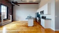 Located in Providence, RI, this single-floor loft space masterfully blends industrial character with modern finishes. The open-concept layout is anchored by rich, warm hardwood flooring that runs throughout, complemented by soaring ceilings adorned with exposed wooden beams, visible ductwork, and a ceiling fan — hallmarks of classic mill-style architecture. The kitchen is a standout feature, offering a central island with a dark stone countertop and integrated sink, white shaker cabinetry, and a full suite of stainless steel appliances including a refrigerator, electric range, and microwave. A stylish sliding barn door adds a rustic-modern touch to the living area. The bedroom showcases original exposed brick walls and large, multi-pane industrial windows that flood the space with natural light and provide views of the surrounding greenery. A wall-mounted mini-split unit ensures year-round comfort. The bathroom is clean and functional, featuring white cabinetry with stone countertops and wood flooring that maintains the cohesive aesthetic throughout. This loft offers a distinctive urban living experience rooted in Providence's storied architectural heritage.