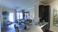 This single-floor residence showcases a thoughtfully designed open-concept layout that seamlessly connects the kitchen, dining, and living areas. The kitchen is equipped with dark espresso cabinetry, a stainless steel sink, and granite-style countertops that extend into a breakfast bar, offering additional seating and a natural transition into the dining space. Crown molding runs throughout, adding a refined architectural touch to the interior.

The living area is generously proportioned, featuring large windows dressed with elegant curtains that allow abundant natural light to fill the room. A ceiling fan adds both style and functionality overhead. The space is centered around a comfortable sofa and a mirrored coffee table, complemented by a bold piece of wall art that adds a vibrant focal point to the room.

Adjacent to the living area, a private bedroom is visible through an open doorway, offering a calm and secluded retreat. The overall color palette is neutral and contemporary, with warm tones that create an inviting ambiance throughout. The open flow between spaces makes the residence feel expansive and well-suited for both everyday living and entertaining.