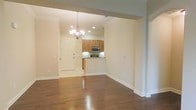 This single-story space showcases an open-concept layout that seamlessly connects the main living area, dining space, and kitchen. Rich, dark hardwood flooring extends throughout the living and dining areas, offering a warm contrast to the light-toned walls and crisp white baseboards and crown molding that frame each room.

The kitchen is positioned at the heart of the space, featuring warm honey-toned wood cabinetry, stainless steel appliances including a microwave and range, and a breakfast bar that separates it from the dining area. Recessed lighting illuminates the kitchen, complemented by a decorative chandelier that anchors the dining space with an elegant touch.

The entryway, finished with light tile flooring, provides a clean transition into the home and is accented by a white paneled door. Recessed lighting throughout ensures consistent, bright illumination across the open floor plan. The neutral wall palette and cohesive design elements create a bright, airy ambiance, making the space feel open and move-in ready.