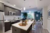 Located at 7480 Birdwood Ave in McLean, VA, this thoughtfully designed 2-bedroom, 2-bathroom residence offers a modern aesthetic with bold design choices throughout. The welcoming entrance hallway features warm wood-tone flooring, a decorative mirror, and a slim console table that set the tone for the stylish interiors beyond.

The open-concept kitchen and living area serves as the heart of the home. The kitchen is outfitted with dark espresso cabinetry, a crisp white quartz waterfall island with bar seating, stainless steel appliances, and tile backsplash — ideal for both everyday cooking and entertaining. Pendant lighting illuminates the island, while recessed ceiling lights add to the bright, modern atmosphere.

The combined dining and living room benefits from a large picture window that draws in natural light and frames leafy outdoor views. Teal accent walls add a vibrant, cohesive design thread throughout the space.

The primary bedroom continues the bold teal accent wall, complemented by natural light from a generous window and its own closet. The second bedroom mirrors this inviting aesthetic. Both bathrooms are finished with dark vanity cabinetry, white quartz countertops, and clean tile work in the shower surround. The residence also includes in-unit laundry and three closets, providing practical storage throughout.
