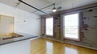 This single-floor space showcases a compelling blend of raw industrial character and modern finishes. The open-plan layout seamlessly connects the kitchen and living areas, creating a versatile, flowing environment. Overhead, painted white wood-plank ceilings are complemented by exposed black metal conduit and pipes, reinforcing the industrial aesthetic throughout. A ceiling fan with a built-in light fixture provides both air circulation and ambient illumination, while pendant lights with delicate glass shades hang above the kitchen peninsula, adding a refined touch.

The kitchen features a granite-topped peninsula with an integrated sink, offering a functional workspace that also serves as a natural divider between the cooking and living zones. Adjacent, the living area benefits from multiple large windows framed by a striking exposed brick accent wall, where layers of paint and aged masonry tell the story of the building's history. These generous windows flood the space with natural light and provide views of the surrounding neighborhood. Polished hardwood flooring runs throughout, tying together the contrasting textures of rough brick and smooth surfaces into a cohesive, characterful interior.