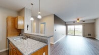 Located at Rivers Pointe in Liverpool, NY, this single-floor apartment offers a well-designed open-concept layout that seamlessly connects the kitchen, dining, and living areas. The kitchen is appointed with warm honey-toned cabinetry, granite countertops, black appliances, and a peninsula with pendant lighting overhead — ideal for casual dining or entertaining. Hardwood-style flooring runs throughout the main living areas, complementing the neutral gray tones of the walls and adding a clean, modern aesthetic. The spacious living room benefits from abundant natural light streaming through a large sliding glass door that opens to a private balcony, offering outdoor views of the surrounding landscape. A ceiling fan enhances comfort in the living space. The bedroom features plush wall-to-wall carpeting and a generously sized window that brings in additional natural light and garden views. The bathroom is well-appointed with a granite-topped vanity with warm wood cabinetry, a full tub-and-shower combination, and hardwood-style flooring that ties the space together. Throughout the unit, clean white trim, ample storage, and a practical single-floor layout provide a comfortable and functional living environment.