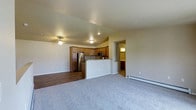 This single-floor space showcases an open-concept design that seamlessly connects the living area and kitchen. The living room is finished with plush neutral carpeting and features a ceiling fan, baseboard heating, and smooth, lightly vaulted ceilings that add a sense of openness. A recessed hallway leads to additional rooms, including what appears to be a bathroom with visible lighting.

The kitchen is well-appointed with warm-toned wood cabinetry, stainless steel appliances including a refrigerator and microwave, and a peninsula-style breakfast bar with a light-colored countertop. The kitchen area transitions to durable hardwood-style flooring, creating a functional and easy-to-maintain surface. Recessed and flush-mount ceiling lighting provides consistent illumination throughout the kitchen and adjoining spaces.

The overall ambiance is clean, practical, and welcoming, with neutral wall tones and thoughtful layout choices that maximize the flow between the living and kitchen areas. The baseboard heater along the living room wall is a notable functional feature, ensuring efficient climate control throughout the space.