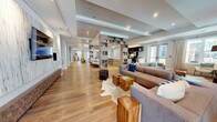 Located at 3420 Legacy Eagle Dr in Mount Pleasant, South Carolina, this single-floor community clubhouse showcases a sophisticated coastal-modern aesthetic that flows seamlessly from one zone to the next. Wide-plank hardwood flooring runs throughout, unifying the expansive open-concept layout beneath coffered and tray ceilings detailed with warm wood accents and recessed lighting.

The main lounge area is generously sized, anchored by large sectional sofas in neutral tones, cowhide rugs, and natural wood stump accent tables. Abundant windows dressed in floor-length linen drapes fill the space with natural light and offer views of the surrounding community. A second lounge zone features a striking teal ceiling detail with a statement ring chandelier, open shelving, and curated artwork, creating a vibrant focal point.

A semi-private nook with built-in bench seating provides a more intimate gathering spot alongside the main social areas. The fully equipped kitchen features dark cabinetry, stainless steel appliances, quartz countertops, and an extended island with bar seating. Adjacent to the kitchen, a large dining table with white upholstered chairs accommodates group gatherings comfortably. Throughout, the design balances refined detail with an approachable, welcoming ambiance.