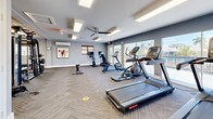 Located at 1900 54th Ave S in Saint Petersburg, Florida, this fitness center offers a generously sized, single-floor exercise space designed to support a full range of workout activities. The room is finished with a neutral gray and white color palette, complemented by patterned low-pile carpet tiles that add texture while maintaining a clean, professional aesthetic.

The space is divided into distinct functional zones. A dedicated cardio area lines the perimeter near large floor-to-ceiling sliding glass doors, which flood the room with natural light and offer views of a neighboring outdoor balcony and palm-lined surroundings. This section houses treadmills, an elliptical trainer, and stationary bikes. Ceiling fans and a wall-mounted mini-split system ensure comfortable climate control throughout.

The strength training zone features a variety of resistance machines with padded seating, a full dumbbell rack, a TRX suspension training wall unit labeled "Training Zone," and a weight bench. A round mirror and small sink station are conveniently integrated into the space. Vibrant framed artwork adorns the walls, adding an energetic and motivating atmosphere.

Recessed rectangular panel lighting provides bright, even illumination throughout the room. An accessible unisex restroom is located on-site, and a clearly marked emergency exit ensures safety compliance. Overall, the space is thoughtfully arranged, clean, and well-maintained.