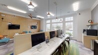 This thoughtfully designed single-floor amenity space blends functionality with contemporary style, offering residents or visitors a welcoming and versatile environment. Large multi-pane windows line the walls throughout, flooding the interior with natural light and providing open views of the surrounding landscaping.

The business center is a standout feature, anchored by a long marble-topped communal desk outfitted with desktop computers and ergonomic seating. Elegant copper-accented pendant lights hang above, adding a refined industrial touch to the workspace. A colorful green-toned area rug grounds the space and adds a pop of personality to the flooring beneath.

Adjacent to the workstation area, a comfortable lounge zone invites relaxation with plush teal accent chairs accented by vibrant orange throw pillows, arranged around small round tables. A textured cork-panel feature wall serves as a backdrop for a wall-mounted flat-screen television, complemented by tasteful sconce lighting. Artwork in ocean-inspired hues adds a creative focal point near the reception-style counter.

The space also connects directly to a fitness area, visible through an interior window, and is accessible via a classic black-framed glass-paned entry door. Crown molding throughout lends an architectural polish, while the eclectic mix of materials and colors creates a modern yet inviting atmosphere.