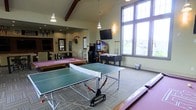 This single-floor game room is designed for recreation and social engagement, offering a well-equipped space filled with a variety of entertainment options. The room's architecture is immediately notable, featuring vaulted ceilings accented by exposed dark wood beams that add warmth and character to the space. Large, floor-to-ceiling windows along one wall flood the interior with abundant natural light, providing views of the surrounding neighborhood while creating an open, airy atmosphere.

The room is outfitted with a full-sized ping pong table at its center, complemented by a classic billiards table finished in a distinctive pink felt. A foosball table and multiple arcade gaming machines offer additional entertainment, while a wall-mounted flat-screen television adds to the recreational options. Pendant lighting and recessed fixtures ensure the space remains well-lit during evening hours.

The flooring features a patterned carpet that defines the gaming area, contributing to the room's comfortable, casual ambiance. Decorative pennant banners and framed artwork on the walls add a lively, communal character. Overall, the space functions as an inviting, activity-rich gathering area suited for group entertainment.