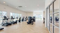 Located at 49 N Assembly Dr in Wilmington, NC, this modern fitness center spans a generous single floor, offering a thoughtfully designed environment for a comprehensive workout experience. The space is divided into two distinct areas: a main gym floor and a separate studio room, both unified by a striking light-toned herringbone hardwood floor that adds warmth and visual interest throughout.

The main gym area is bright and open, featuring high ceilings, recessed lighting, and expansive windows along multiple walls that flood the space with natural light and provide views of the surrounding community. The room is well-appointed with a full suite of True-brand strength machines, cable systems, treadmills, ellipticals, stationary bikes, and a dedicated dumbbell rack. A wall-mounted flat-screen TV adds convenience for users during workouts.

The adjacent studio room offers a more versatile, open-plan layout beneath a warm wood-paneled ceiling with recessed lighting. A full-panel glass garage door opens to the outdoors, allowing fresh air and additional light to flow in. The studio is stocked with yoga mats, stability balls, resistance equipment, and other group fitness accessories, making it ideal for stretching, yoga, or guided classes. Together, the two areas create a complete, modern fitness facility suited for a wide range of exercise routines.