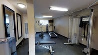 This fitness room is a dedicated exercise space designed to support a variety of workout routines. The room features dark rubber flooring, which provides both durability and traction underfoot, and is finished with textured ceilings and neutral-toned walls that give the space a clean, functional aesthetic. Fluorescent lighting fixtures illuminate the room evenly, ensuring good visibility throughout.

The cardio section includes two treadmills and an upright stationary bike, catering to users with varying cardiovascular training preferences. A dumbbell rack with a selection of free weights is positioned along the wall, providing options for strength and resistance training. An adjustable weight bench is available for use with the free weights. On the opposite side of the room, a cable pulley machine offers additional strength-training versatility for a range of upper and lower body exercises.

A wall-mounted flat-screen television is positioned near the front of the room, allowing users to stay entertained during workouts. A window fitted with vertical blinds introduces a degree of natural light. A paper towel or sanitary wipe dispenser is conveniently accessible near the entrance, supporting cleanliness and equipment maintenance.