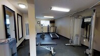 This fitness room is a dedicated exercise space designed to support a variety of workout routines. The room features dark rubber flooring, which provides both durability and traction underfoot, and is finished with textured ceilings and neutral-toned walls that give the space a clean, functional aesthetic. Fluorescent lighting fixtures illuminate the room evenly, ensuring good visibility throughout.

The cardio section includes two treadmills and an upright stationary bike, catering to users with varying cardiovascular training preferences. A dumbbell rack with a selection of free weights is positioned along the wall, providing options for strength and resistance training. An adjustable weight bench is available for use with the free weights. On the opposite side of the room, a cable pulley machine offers additional strength-training versatility for a range of upper and lower body exercises.

A wall-mounted flat-screen television is positioned near the front of the room, allowing users to stay entertained during workouts. A window fitted with vertical blinds introduces a degree of natural light. A paper towel or sanitary wipe dispenser is conveniently accessible near the entrance, supporting cleanliness and equipment maintenance.