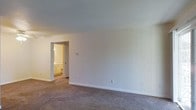 This single-story space presents a straightforward, functional layout centered around a spacious main room finished with wall-to-wall carpet in a neutral gray-brown tone. The room features clean white walls and a smooth white ceiling, creating a bright and open atmosphere. Multiple electrical outlets are positioned along the baseboards, offering practical flexibility for furniture arrangement. A doorway leads directly into an adjoining bathroom, which is visible from the main room and features warm-toned cabinetry, suggesting a vanity and toilet within a compact but functional layout. The bathroom's warmer lighting contrasts with the cooler, even illumination of the main room. The overall design is minimal and unfurnished, making it easy to envision a variety of uses for the space. With its neutral color palette, carpeted flooring, and accessible bathroom, this space offers a clean, move-in-ready environment suited for a range of occupants.