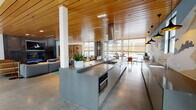 Located at 108 Moffett Blvd in Mountain View, CA, this single-story community clubhouse showcases a sophisticated, open-plan design that seamlessly integrates multiple functional zones. Polished concrete flooring flows throughout, complemented by striking slatted wood ceilings that lend warmth and architectural continuity across the entire space.

The centerpiece is a fully equipped communal kitchen featuring an expansive stainless steel island with a built-in gas cooktop, range hood, and bar seating. A vivid mosaic tile mural depicting a mountainous landscape serves as a dramatic backsplash, adding an artistic focal point. Pendant lighting and abundant counter space complete this well-appointed area.

Adjacent to the kitchen, a casual dining zone offers multiple wood-and-chair table configurations beneath woven roller shades that filter generous natural light from floor-to-ceiling windows. Two distinct lounge areas provide varied seating arrangements — one anchored by a bold yellow-green area rug with contemporary sofas, curated textile wall art, and a custom wooden shelving unit; the other centered on a built-in fireplace framed in dark metal paneling with a wall-mounted TV.

A bright corridor lined with large glass sliding doors opens directly onto an outdoor courtyard, bringing the surrounding residential community into view. Thoughtful decorative accents, including fiber art wall hangings and natural wood details, contribute to a warm, curated ambiance throughout.