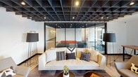 This thoughtfully designed single-floor space blends contemporary aesthetics with functional versatility across several distinct zones. The layout, as revealed in the dollhouse view, encompasses a generous open-plan area that transitions seamlessly between a lounge, dining, and collaborative workspace.

A dramatic dark grid-panel ceiling anchors the central lounge area, creating a bold architectural statement overhead. A plush, neutral-toned sofa and accent chairs are arranged around a coffee table, while a full-length shuffleboard table adds a recreational element nearby. Floor lamps with oversized shades provide warm ambient lighting throughout.

A glass-walled enclosure with a black steel frame defines a semi-private meeting or presentation room, visible from the lounge and furnished with tiered seating. This transparent partition maintains visual connectivity while offering acoustic separation.

The dining area features light wood tables and upholstered chairs arranged around a fireplace topped with a wall-mounted flat-screen display, creating a warm and inviting gathering spot. Adjacent wall art adds a curated touch.

The kitchenette is clean and functional, finished with white cabinetry, matte black hardware, a stainless steel sink, and light wood-tone flooring that carries throughout the space, tying all zones together cohesively.