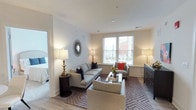 This single-floor residence offers a thoughtfully designed living space that balances comfort and contemporary style. The living room serves as the heart of the home, featuring light-toned hardwood flooring and neutral white walls that create a clean, airy atmosphere. Natural light streams in through large windows fitted with white blinds, offering pleasant views of the surrounding neighborhood and enhancing the room's bright, welcoming feel.

The living area is generously proportioned, accommodating a full seating arrangement that includes a light gray sofa adorned with navy accent pillows, a coordinating armchair with a bold red cushion, and an additional upholstered chair. A glass-top coffee table anchors the seating group atop a geometric-patterned area rug in warm neutral tones. Ambient lighting is provided by elegantly styled table lamps, while a decorative sunburst mirror adds visual interest to one wall.

A dark wood dresser along one wall provides functional storage, and an open doorway reveals an adjoining bedroom, suggesting a well-connected, flowing floor plan. The overall ambiance is polished, calm, and move-in ready.