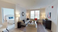 This single-floor residence offers a thoughtfully designed living space that balances comfort and contemporary style. The living room serves as the heart of the home, featuring light-toned hardwood flooring and neutral white walls that create a clean, airy atmosphere. Natural light streams in through large windows fitted with white blinds, offering pleasant views of the surrounding neighborhood and enhancing the room's bright, welcoming feel.

The living area is generously proportioned, accommodating a full seating arrangement that includes a light gray sofa adorned with navy accent pillows, a coordinating armchair with a bold red cushion, and an additional upholstered chair. A glass-top coffee table anchors the seating group atop a geometric-patterned area rug in warm neutral tones. Ambient lighting is provided by elegantly styled table lamps, while a decorative sunburst mirror adds visual interest to one wall.

A dark wood dresser along one wall provides functional storage, and an open doorway reveals an adjoining bedroom, suggesting a well-connected, flowing floor plan. The overall ambiance is polished, calm, and move-in ready.