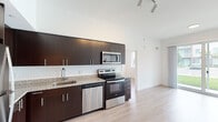 This single-floor unit showcases a contemporary design with a cohesive aesthetic throughout. The kitchen is a standout feature, equipped with a full suite of stainless steel appliances including a range, microwave, dishwasher, and refrigerator. Rich dark-toned cabinetry provides ample storage, complemented by granite countertops and a classic white subway tile backsplash. Soaring ceilings and track lighting lend the kitchen an open, airy quality that extends into the adjacent living area. Light-toned hardwood-style flooring flows throughout the main living spaces, enhancing the bright, clean atmosphere created by crisp white walls.

The living area benefits from large windows fitted with horizontal blinds, offering views of palm trees and the surrounding streetscape, allowing natural light to fill the room. Two bedrooms feature the same light flooring and well-sized windows with outdoor views.

Both bathrooms maintain the unit's consistent design language, each featuring granite-topped dark vanities, large mirrors, tile flooring, and a soaker tub with a tiled surround. The overall layout, as seen in the floorplan view, is efficiently organized, offering comfortable and functional living spaces throughout.