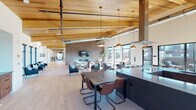 This two-story space is defined by its striking architectural character, most notably the vaulted wood-plank ceiling supported by exposed timber beams that crowns the main level. Light hardwood flooring runs throughout, complementing the warm tones of the natural wood overhead and creating a cohesive, contemporary aesthetic.

The main floor opens into a generously scaled communal area that integrates dining and lounge functions seamlessly. A dark, streamlined kitchen island anchors one side of the space, paired with a full kitchenette featuring stainless steel appliances, wood cabinetry, and a herringbone tile backsplash. Pendant lighting in brushed brass and copper tones adds warmth above dining tables and gathering areas. A textured accent wall houses a fireplace and mounted television, creating a comfortable lounge setting surrounded by large windows that invite abundant natural light.

A distinctive oval-framed nook provides an intimate seating alcove, while glass-walled rooms offer flexible conference and meeting space, including a formal conference room adorned with a vintage railroad map mural.

The second floor, accessed via a modern staircase with black metal railings, features a game room with a billiards table, a linear fireplace set against slate-toned wall panels, and banquette seating with amber sconce lighting. Outdoor views are accessible from multiple vantage points throughout both levels.