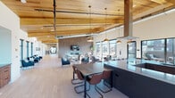 This two-story space is defined by its striking architectural character, most notably the vaulted wood-plank ceiling supported by exposed timber beams that crowns the main level. Light hardwood flooring runs throughout, complementing the warm tones of the natural wood overhead and creating a cohesive, contemporary aesthetic.

The main floor opens into a generously scaled communal area that integrates dining and lounge functions seamlessly. A dark, streamlined kitchen island anchors one side of the space, paired with a full kitchenette featuring stainless steel appliances, wood cabinetry, and a herringbone tile backsplash. Pendant lighting in brushed brass and copper tones adds warmth above dining tables and gathering areas. A textured accent wall houses a fireplace and mounted television, creating a comfortable lounge setting surrounded by large windows that invite abundant natural light.

A distinctive oval-framed nook provides an intimate seating alcove, while glass-walled rooms offer flexible conference and meeting space, including a formal conference room adorned with a vintage railroad map mural.

The second floor, accessed via a modern staircase with black metal railings, features a game room with a billiards table, a linear fireplace set against slate-toned wall panels, and banquette seating with amber sconce lighting. Outdoor views are accessible from multiple vantage points throughout both levels.