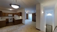 This single-floor space showcases an open-concept design that seamlessly connects the kitchen and living areas. The kitchen is outfitted with warm, dark wood cabinetry that extends to the upper walls, offering generous storage alongside light-toned countertops and a white refrigerator. A raised breakfast bar peninsula serves as a natural divider between the kitchen and the carpeted living space, adding both functionality and definition to the layout.

The living area features neutral, light-toned carpeting that extends throughout, contributing to a cohesive and comfortable feel across the space. A ceiling fan with an integrated light fixture is centrally positioned, providing both air circulation and ambient lighting. Wall-mounted sconce lighting adds supplemental illumination along the walls.

Vertical blinds on a sliding glass door at the far end of the living area allow for adjustable natural light and hint at outdoor access. Interior doors with wood-toned finishes contribute to the warm, consistent aesthetic found throughout. The layout is practical and straightforward, with clear sightlines from the kitchen through to the living area, making the space feel open and connected.