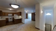 This single-floor space showcases an open-concept design that seamlessly connects the kitchen and living areas. The kitchen is outfitted with warm, dark wood cabinetry that extends to the upper walls, offering generous storage alongside light-toned countertops and a white refrigerator. A raised breakfast bar peninsula serves as a natural divider between the kitchen and the carpeted living space, adding both functionality and definition to the layout.

The living area features neutral, light-toned carpeting that extends throughout, contributing to a cohesive and comfortable feel across the space. A ceiling fan with an integrated light fixture is centrally positioned, providing both air circulation and ambient lighting. Wall-mounted sconce lighting adds supplemental illumination along the walls.

Vertical blinds on a sliding glass door at the far end of the living area allow for adjustable natural light and hint at outdoor access. Interior doors with wood-toned finishes contribute to the warm, consistent aesthetic found throughout. The layout is practical and straightforward, with clear sightlines from the kitchen through to the living area, making the space feel open and connected.