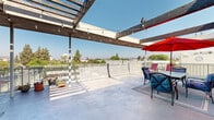 Located at 510 E Third St in Pomona, California, this rooftop terrace offers a compelling outdoor experience with sweeping panoramic views of the surrounding cityscape, lush tree canopy, and distant mountain ranges. The open-air deck features a polished concrete floor that extends generously across the rooftop, providing ample space for multiple functional zones.

A slatted wood pergola spans a significant portion of the terrace, draped with café-style string lights that lend a warm, inviting ambiance during evening gatherings. Beneath the pergola, a dedicated dining area is anchored by a patio table and chairs shaded by a bold red market umbrella, making it ideal for al fresco meals. A perimeter safety railing with a sleek metal mesh design frames the terrace, maintaining unobstructed sightlines to the horizon.

A separate lounge zone features a generously sized sectional sofa with vibrant red cushions set against a striking mosaic tile accent wall, creating a cozy, semi-sheltered retreat. Additional seating and potted plants are thoughtfully placed throughout, adding greenery and character to the space.

This rooftop terrace is well-suited for social gatherings, events, or relaxed outdoor enjoyment, combining functional design with open-sky exposure and notable architectural detail.