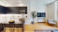 Located at 1401 Spruce St in the heart of Philadelphia, this thoughtfully designed single-floor residence combines contemporary style with refined details throughout. Light hardwood flooring flows seamlessly through the open-plan living and kitchen areas, anchored by large windows that invite natural light and offer urban views of the surrounding cityscape.

The kitchen is a standout feature, showcasing a two-tone cabinetry scheme with crisp white lower cabinets and dark upper cabinets, complemented by marble-look quartz countertops and backsplash, brushed gold hardware, and stainless steel appliances. A central island with bar seating connects the kitchen to the living area, where a tufted navy blue velvet sofa and curated artwork create an inviting atmosphere.

The bedroom is elegantly appointed with a textured charcoal accent wall, recessed lighting, and a window with city views, lending a calm and sophisticated ambiance. A hallway with warm recessed lighting, a large round gold-framed mirror, and a frosted glass door leads to additional spaces, including a dart board recreation nook and a dedicated game room featuring bold Art Deco-inspired wallpaper in black and gold. A powder room with hexagonal patterned tile flooring and gold fixtures adds a polished finishing touch.