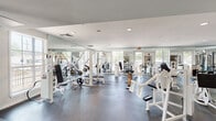 This fitness center in New Orleans, Louisiana, is a spacious, single-floor facility thoughtfully organized into distinct workout zones. The interior features a clean, bright aesthetic with white walls, recessed lighting, and large windows that invite natural light throughout the space.

The cardio area is lined with a robust selection of treadmills, ellipticals, and stair climbers arranged in orderly rows, complemented by wall-mounted televisions to keep members engaged during workouts. Durable wood-tone and carpeted flooring define this section, offering comfort underfoot.

The strength training areas are expansive and well-appointed, featuring multiple cable machines, weight benches, squat racks, free weights, and a full dumbbell rack. Floor-to-ceiling mirrors line the walls, enhancing the sense of openness while allowing members to monitor their form. Rubber flooring throughout these zones provides safety and stability.

A dedicated studio room offers cycling bikes, ellipticals, and additional cardio equipment, alongside a wall-mounted TV and built-in cabinetry, creating an intimate setting for focused workouts. Structural columns and defined corridors connect the various zones seamlessly, contributing to an organized flow throughout the facility. The overall ambiance is clean, modern, and professional.