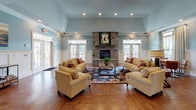 Located at Amelia Station in Clayton, NC, this single-story clubhouse offers a well-appointed community gathering space designed with classic Southern charm. The centerpiece of the main lounge is a floor-to-ceiling stacked stone fireplace, flanked by multiple sets of French doors that flood the room with natural light and provide views of the outdoor surroundings. Soaring vaulted ceilings, soft blue walls, white wainscoting with crown molding, and rich hardwood flooring give the space a warm, refined character. Comfortable seating arrangements with plush sofas and accent chairs surround the fireplace atop a vibrant area rug, creating an inviting atmosphere for relaxation and socializing. Adjacent to the lounge is a dining nook featuring a round wooden table, leather arm chairs, a wall-mounted flat-screen TV, and a built-in bookcase — ideal for casual meetings or quiet reading. The fully equipped kitchen boasts rich dark cabinetry, dark granite countertops, stainless steel appliances including a side-by-side refrigerator, a double-basin sink with a generous island, and a coffee station. A game room with a billiard table and fitness equipment rounds out the amenities, making this clubhouse a comprehensive hub for community life.