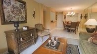 This single-floor residence exudes a warm, classic elegance throughout its thoughtfully designed interior. The living area sets a refined tone with rich hardwood furnishings, including an ornate wooden dresser and a traditionally upholstered armchair that anchor the space with timeless character. A decorative area rug defines the seating zone, while an artistic wall piece and a candle sconce add curated charm to the golden-hued walls. Light-toned tile flooring flows seamlessly across the open layout, lending a bright and cohesive feel. A wooden coffee table with an inlaid tray display serves as an elegant centerpiece, making the living area ideal for both relaxed gatherings and stylish entertaining. The space opens naturally into a formal dining area, where a classic chandelier casts warm, inviting light over a substantial wooden dining table surrounded by matching upholstered chairs. A glimpse into the adjacent kitchen area reveals a bright and accessible layout. The overall ambiance is one of traditional sophistication, with warm earth-tone walls, carefully chosen décor, and a consistent design language that creates a welcoming and well-appointed atmosphere throughout the home.