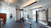 This fitness center offers a comprehensive workout environment thoughtfully designed with both functionality and comfort in mind. The space is organized into distinct exercise zones, each outfitted with professional-grade equipment. The strength training area features Hoist cable machines, a functional trainer, weight benches, a rack of dumbbells, and stability balls, providing a full range of resistance training options. Adjacent to this, a dedicated cardio zone lines the perimeter wall, where treadmills and elliptical machines are positioned directly in front of expansive floor-to-ceiling windows, allowing users to exercise while taking in panoramic views of the surrounding cityscape and treetops.

The flooring throughout is covered in durable dark rubber tile, providing both safety and sound absorption. Overhead, linear pendant lighting and recessed fixtures illuminate the space evenly, complemented by abundant natural light streaming through the large windows. A warm-toned wood built-in shelving and storage unit anchors one end of the facility, alongside a small service counter area. The clean white walls and modern architectural details contribute to an organized, professional atmosphere suited for focused, effective training.