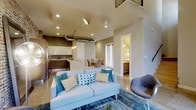 This contemporary 2-floor space makes a striking impression with its soaring ceilings and open-concept design. The main living area is bright and inviting, anchored by a spacious lounge area with room for comfortable seating and a sleek coffee table. An industrial-style exposed duct adds a modern architectural accent overhead, complementing the recessed lighting that illuminates the space throughout. The open-plan kitchen flows seamlessly from the living area, featuring crisp white upper cabinetry, dark lower cabinetry, a light-toned countertop island, stainless steel appliances, and pendant lighting overhead. Adjacent to the kitchen, a dedicated dining area offers natural light through a slatted window blind. The neutral warm-toned walls and polished concrete-style flooring create a cohesive, sophisticated palette that runs throughout the main level. A second floor is accessible and visible from the main living area via an upper-level overlook railing, adding architectural depth and vertical drama. A hallway off the main living space leads to additional rooms, including what appears to be a bathroom with warm lighting. The overall ambiance is modern, clean, and well-appointed, with thoughtful design details that balance style and functionality.