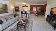 This single-floor residence showcases an open-concept design that thoughtfully integrates the living, dining, and kitchen areas into one cohesive and inviting space. The living area is anchored by plush carpeting layered with an ornate area rug, complementing a neutral-toned sofa and a glass-top coffee table that adds a refined touch. Recessed lighting and crown molding along the ceiling contribute a polished, finished look throughout.

A raised breakfast bar with dark leather bar stools serves as a natural transition between the living space and the kitchen, which features white cabinetry, under-cabinet lighting, and open shelving above for decorative accents. The kitchen's clean, bright aesthetic contrasts elegantly with the bold deep-red accent wall in the adjacent dining area.

The dining space is characterized by its warm, dramatic wall color, framed artwork, and a classically styled dining set that creates an intimate atmosphere. Lush indoor greenery is thoughtfully placed throughout, adding life and vibrancy. Overall, the space balances comfort and sophistication through its cohesive color palette and carefully curated design elements.