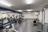 Located at 450 Victoria Ave in Windsor, Ontario, this fitness facility offers a comprehensive, single-floor gym designed to support a wide range of workout routines. The space features durable dark rubber flooring throughout the main exercise area, providing both safety and functionality for various training activities.

The gym is divided into distinct workout zones. The cardio section is well-equipped with multiple treadmills, elliptical trainers, and stationary bikes, all arranged along a full-length mirrored wall accented with navy blue trim, which enhances the sense of space and allows users to monitor their form. The strength training area includes a cable machine, weight bench, barbell with plate weights, a power tower, and additional resistance equipment, offering a thorough selection for muscle conditioning.

A dedicated free weights and accessories corner features stability balls, a dumbbell rack, and benches. An adjacent utility area with tile flooring houses a drinking fountain, a storage counter, and a hand sanitizer station, supporting hygiene and convenience. Overhead, fluorescent strip lighting ensures bright, even illumination throughout. Exposed ductwork along the ceiling adds an industrial character to the space. The overall layout is open and organized, providing an accessible and functional environment for a variety of fitness goals.