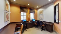 This single-floor business center offers a functional and welcoming environment designed for focused work. The room features warm amber-toned accent walls complemented by crisp white upper walls and dark wood trim, creating a polished, professional atmosphere. Multiple computer workstations are arranged along a built-in dark wood desk that runs along the perimeter, each equipped with a desktop monitor and comfortable task chair with upholstered seating. Two windows dressed with layered Roman shades allow natural light to filter into the space while maintaining privacy, contributing to a bright yet comfortable ambiance. Recessed ceiling lighting supplements the natural light, ensuring consistent illumination throughout. Decorative botanical artwork, featuring coral and seaweed motifs in neutral tones, adorns the white wall, adding a refined touch to the space. The neutral carpeted flooring ties the design elements together cohesively. Overall, this business center strikes a balance between utility and comfort, providing an inviting setting suited for work, browsing, or printing needs.