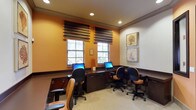 This single-floor business center offers a functional and welcoming environment designed for focused work. The room features warm amber-toned accent walls complemented by crisp white upper walls and dark wood trim, creating a polished, professional atmosphere. Multiple computer workstations are arranged along a built-in dark wood desk that runs along the perimeter, each equipped with a desktop monitor and comfortable task chair with upholstered seating. Two windows dressed with layered Roman shades allow natural light to filter into the space while maintaining privacy, contributing to a bright yet comfortable ambiance. Recessed ceiling lighting supplements the natural light, ensuring consistent illumination throughout. Decorative botanical artwork, featuring coral and seaweed motifs in neutral tones, adorns the white wall, adding a refined touch to the space. The neutral carpeted flooring ties the design elements together cohesively. Overall, this business center strikes a balance between utility and comfort, providing an inviting setting suited for work, browsing, or printing needs.