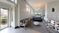 This elegantly appointed residential lobby makes a striking first impression with its soaring double-height ceilings and refined Art Deco-inspired architecture. The space is bathed in a bright, neutral palette of crisp whites and soft grays, enhanced by natural light filtering through clerestory windows positioned high along the walls.

The lounge area is thoughtfully furnished with a sleek black leather sofa, geometric patterned accent chairs with chrome frames, and a metallic pouf ottoman, all anchored by a plush area rug. A dramatic sculptural chandelier with candelabra-style arms serves as a focal point overhead, while a floor-to-ceiling mirror wall amplifies the sense of depth and grandeur throughout the seating area.

A grand staircase with warm-toned stone steps and polished chrome railings rises from the lobby level, flanked by glass block architectural details that add a distinctive period character. The lower level features a dedicated mailroom wall lined with rows of individual resident mailboxes, illuminated by warm sconce lighting for easy access.

The lobby's generous proportions, clean architectural lines, crown molding, and carefully curated furnishings combine to create a welcoming yet sophisticated atmosphere that sets an elevated tone for the entire building.