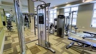 This fitness center is designed to support a comprehensive workout experience, offering a thoughtfully arranged selection of strength training and exercise equipment. The space is outfitted with multiple cable machines, a Precor strength station, and additional resistance equipment, providing options for a full-body training routine. The layout allows for comfortable movement between stations without feeling cramped.

The flooring features a patterned carpet in neutral earth tones, lending a clean and functional aesthetic to the space. Recessed lighting in the ceiling provides consistent, even illumination throughout, while large windows along one wall invite natural light and offer views of the surrounding exterior, contributing to an open and energizing atmosphere.

The walls are finished in a warm neutral tone, complementing the overall design and keeping the focus on the equipment. Mirrors are visible along portions of the walls, a practical feature that supports proper form during exercise while also adding a sense of depth to the room. Overall, the fitness center is well-maintained and organized, making it a practical and inviting environment for exercise.