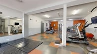 Located at 1100 Everton Pl in Riverside, California, this well-equipped fitness center offers a comprehensive workout environment across a single open floor. The space is defined by a bold, energetic color scheme featuring vibrant orange accent walls paired with two-tone gray and white wainscoting, creating a motivating atmosphere throughout. The distinctive checkered flooring in orange and gray tiles adds to the dynamic aesthetic while providing a durable, easy-to-maintain surface.

The layout is thoughtfully organized to accommodate a variety of workout styles. A dedicated cardio zone houses multiple treadmills and stationary bikes, each positioned for comfortable use. A strength training area features a multi-station cable machine alongside a dumbbell rack with a full range of free weights and an adjustable weight bench. Large wall-mounted mirrors expand the sense of space and allow for form monitoring during exercise. Padded floor mats are available for stretching, yoga, and bodyweight training.

Structural columns define zones within the open floor plan without disrupting the flow of the space. Overhead fluorescent lighting ensures the entire facility is evenly and brightly illuminated. Large glass doors along one wall fill the entry area with natural light and provide views of the outdoor courtyard beyond, creating a welcoming and airy feel. A wall-mounted television adds entertainment for cardio sessions, rounding out this fully functional community fitness facility.