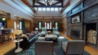 Located at 5550 Bexley Way NW in Concord, NC, this single-floor community clubhouse is a well-appointed gathering space designed for comfort and socializing. The main lounge area commands immediate attention with its dramatically high ceilings, exposed wood beam detailing, and a striking coffered ceiling section that adds architectural depth. A floor-to-ceiling stacked stone fireplace serves as a central focal point, flanked by a mounted flat-screen TV, and surrounded by an inviting mix of leather and upholstered seating arrangements set atop patterned area rugs over warm hardwood floors. Teal drapery panels and vibrant accent colors throughout lend the space a lively, modern character. Clerestory windows flood the room with abundant natural light. A dedicated game area features a full-sized billiards table, while a separate lounge zone offers a large sectional sofa, built-in media cabinetry, and a coffered ceiling with refined trim work. The kitchen is outfitted with rich dark wood cabinetry, granite countertops, stainless steel appliances including a full-sized refrigerator, microwave, and dishwasher, and tile flooring — ideal for catered events or community gatherings. The overall ambiance is warm, functional, and welcoming.