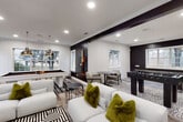 This single-floor amenity space is designed with a bold, contemporary aesthetic that blends entertainment and comfort seamlessly. The color palette centers on vibrant green accent walls, crisp white, and dramatic black trim, creating a visually striking atmosphere throughout. Light wood-look flooring and recessed lighting run consistently across the space, tying the various zones together cohesively.

The main lounge area is generously sized, featuring plush white sofas accented with olive-green throw pillows, a marble-top coffee table, and bold black-and-white geometric area rugs. A wall-mounted flat-screen TV and textured patterned wallpaper add visual interest to the seating zone. Large windows allow ample natural light to fill the room, offering views of the surrounding greenery.

The game room section is a standout feature, equipped with a sleek white pool table illuminated by two pendant gold dome lights, a foosball table, and additional lounge seating arranged around a small bistro table — ideal for casual gatherings.

A dedicated coffee bar station serves as a refined refresh point, complete with dark countertops, gray cabinetry, a built-in beverage refrigerator, a Keurig coffee maker, and a bold black-and-white hexagonal tile backsplash. Complimentary snacks and beverages are thoughtfully displayed, adding a welcoming, hospitality-oriented touch to the overall space.