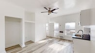 Located at 744 Beacon Ave in the heart of Los Angeles, CA, this single-floor unit offers a clean, modern interior with a cohesive, minimalist design throughout. The open living area is bathed in natural light streaming through multiple windows fitted with blinds, highlighting the light-toned wood-look flooring that runs seamlessly across the entire space. A stylish ceiling fan with a brushed gold accent anchors the main living area, which features notable ceiling height that adds a sense of openness. Built-in open shelving and closet niches along one wall provide convenient storage solutions without interrupting the clean lines of the space. A wall-mounted heater unit is positioned near the entrance for efficient climate control. The kitchen area is well-appointed with crisp white cabinetry, a dark countertop, a stainless steel sink, and a bold dark subway tile backsplash that adds a striking contrast to the bright surroundings. The bathroom maintains the same light, airy aesthetic with all-white finishes, a pedestal sink, a mirrored medicine cabinet, and a window that brings in natural light while offering a view of an adjacent brick wall, adding a touch of urban character. Overall, the space presents a refreshed, move-in-ready interior well-suited for urban living in Los Angeles.