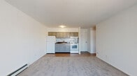 This single-floor residential unit offers a practical and functional layout designed for comfortable everyday living. The open-plan living and kitchen area forms the heart of the space, with the kitchen situated along one wall and finished with gray-toned cabinetry, a marble-style tile backsplash, a white refrigerator, an electric range with oven, and a single-basin sink. The kitchen transitions seamlessly into the carpeted living area, which benefits from a large sliding glass door fitted with vertical blinds, allowing generous natural light to fill the room. A wall-mounted air conditioning unit and baseboard heating are present, ensuring year-round climate control.

The bedroom is a well-proportioned, carpeted room featuring a standard window that brings in additional natural light and outdoor views, along with baseboard heating. A double sliding-door closet provides ample storage space for clothing and personal belongings.

The bathroom is accessed via a narrow hallway and features tile flooring, a toilet, a vanity with storage, and a full bathtub with a shower and curtain rod. The clean, neutral palette throughout the unit — white walls and light-toned finishes — creates a bright and airy atmosphere. Hardwood-style flooring in the kitchen area contrasts with the soft carpeting found in the living and sleeping spaces.