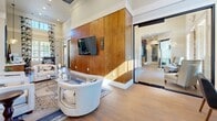 Located at 1035 Scales Rd in Suwanee, GA, this single-floor space offers a thoughtfully designed open layout combining a living room, dining room, and kitchen in a cohesive, stylish environment. The living room commands attention with soaring ceilings, a dramatic floor-to-ceiling wood-paneled accent wall with a wall-mounted flat-screen TV, and flanking wall sconces. Comfortable seating is arranged around a glass-and-gold coffee table atop a patterned area rug, while large windows dressed in bold, marbled blue-and-white drapes invite generous natural light. Wood-look tile flooring flows consistently throughout the space, adding warmth and continuity. The kitchen showcases mid-century modern sensibilities with rich warm-toned wood cabinetry, open floating shelves, a vibrant aqua tile backsplash, white countertops, and a mosaic-tiled island. Stainless steel appliances, including a double wall oven, dishwasher, and refrigerator, complete the functional and visually striking space. The dining area features multiple seating arrangements with round tables, leather-upholstered chairs, and the same bold drapery that ties the design together. Decorative sculptural accents and a sunburst mirror add artistic flair throughout, giving this space a curated, design-forward character.