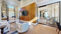 Located at 1035 Scales Rd in Suwanee, GA, this single-floor space offers a thoughtfully designed open layout combining a living room, dining room, and kitchen in a cohesive, stylish environment. The living room commands attention with soaring ceilings, a dramatic floor-to-ceiling wood-paneled accent wall with a wall-mounted flat-screen TV, and flanking wall sconces. Comfortable seating is arranged around a glass-and-gold coffee table atop a patterned area rug, while large windows dressed in bold, marbled blue-and-white drapes invite generous natural light. Wood-look tile flooring flows consistently throughout the space, adding warmth and continuity. The kitchen showcases mid-century modern sensibilities with rich warm-toned wood cabinetry, open floating shelves, a vibrant aqua tile backsplash, white countertops, and a mosaic-tiled island. Stainless steel appliances, including a double wall oven, dishwasher, and refrigerator, complete the functional and visually striking space. The dining area features multiple seating arrangements with round tables, leather-upholstered chairs, and the same bold drapery that ties the design together. Decorative sculptural accents and a sunburst mirror add artistic flair throughout, giving this space a curated, design-forward character.