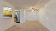 This single-floor unit offers a practical and well-organized layout across several connected living spaces. The open living area is generously sized, featuring neutral wall-to-wall carpeting, a ceiling fan with integrated lighting, and a baseboard heating system. A wall-mounted air conditioning unit provides additional climate control, and a window brings in natural light with views of surrounding greenery.

The kitchen is accessible via a pass-through opening from the living area, offering a functional workspace with light wood cabinetry, dark countertops, and tile flooring. Stainless steel appliances include a gas range, over-the-range microwave, dishwasher, and refrigerator, providing a complete set of modern conveniences.

A second carpeted room, suitable as a bedroom, offers a quiet and neutral space with ample room for furnishings. This room also houses a dedicated laundry closet containing a stacked washer and dryer unit, adding significant convenience.

The bathroom is finished with large-format neutral tiles on both the floor and the tub surround, complemented by a white soaking tub, toilet, and a dark wood vanity with a white countertop. Baseboard heating is also present in this room. Overall, the unit maintains a clean, functional aesthetic throughout.
