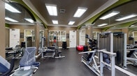 The Ibis Reserve Fitness Center in West Palm Beach, Florida, is a thoughtfully designed workout facility that provides residents with a comprehensive range of exercise options. The space features a distinctive interior aesthetic, highlighted by warm taupe walls accented with bold green arched details, white crown molding, and a rubber-floored layout that prioritizes both safety and function.

The cardio zone is equipped with multiple treadmills and an elliptical trainer, positioned alongside large windows that frame views of lush Florida palm trees, filling the area with natural light and a refreshing outdoor connection. A wall-mounted television accompanies the cardio machines for entertainment during workouts.

The strength training area is thoroughly outfitted with Precor selectorized machines, a multi-functional cable station, a weight bench, and a well-stocked dumbbell rack covering a range of weights. Exercise balls and additional accessories are available for functional and flexibility training. Full-length mirrors line one wall, enhancing the sense of space and allowing users to monitor their form throughout workouts.

Overhead, recessed panel lighting ensures the entire facility is evenly and brightly illuminated. The space is clean, organized, and efficiently arranged to accommodate a variety of fitness routines, making it a valuable amenity for residents of the Ibis Reserve community.