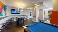 Located at 540 Selma St in Savannah, Georgia, this single-floor community amenity space offers a dynamic and well-appointed environment designed for both recreation and relaxation. The open-plan layout is anchored by a spacious game room featuring a blue felt pool table, foosball table, and a wall-mounted flat-screen TV, all illuminated by a striking modern chandelier and recessed lighting. Dark hardwood-style flooring runs throughout, adding warmth and cohesion to the space. Bold, vibrant orange modular seating units with colorful patterned cushions serve as eye-catching lounge furniture in the central gathering area, complemented by contemporary artwork adorning the white walls. Natural light filters in through large windows, brightening the lounge zone. A dedicated kitchenette area is outfitted with stainless steel appliances including a full-size side-by-side refrigerator with ice and water dispenser, a dishwasher, a wall-mounted TV, and ample white cabinetry with granite-style countertops. A separate work or service nook provides additional counter space, cabinetry, and printing equipment. Recessed ceiling lighting throughout ensures a bright, welcoming atmosphere across all areas of the space.