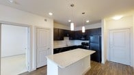 This single-floor residence showcases a thoughtfully designed open-concept layout that centers around a striking kitchen. The kitchen is outfitted with rich, dark espresso cabinetry that extends to the upper walls, providing ample storage while creating a bold visual contrast against the light-toned walls. A generously sized central island features a speckled granite countertop with a gracefully curved edge, offering additional workspace and serving as a natural gathering point. Overhead, two sleek cylindrical pendant lights hang at varying heights, casting warm, focused illumination over the island. Recessed lighting throughout the ceiling further enhances the bright, welcoming ambiance. The kitchen is fully equipped with black appliances, including a refrigerator, range, and built-in microwave, maintaining a cohesive, contemporary aesthetic. A stainless steel sink is positioned along the perimeter cabinetry beneath a window. The flooring features wood-look planks that lend warmth and continuity across the space. A clean white interior door provides access to an adjacent room, reinforcing the crisp, modern palette. The overall design balances functionality with style, creating a polished and practical living environment.