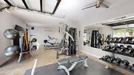 Located in Scottsdale, Arizona, this single-room fitness center offers a well-organized and fully equipped workout space designed to support a wide range of exercise routines. The interior features polished concrete flooring throughout, with rubber mat sections strategically placed beneath the primary equipment zones for added safety and comfort. Recessed lighting and a ceiling fan keep the space bright and well-ventilated, while a wall-mounted mini-split air conditioning unit ensures a comfortable environment year-round. A large roll-up garage door opens the space to natural light and fresh air, lending an open, airy quality to the room. The fitness equipment is comprehensive and includes a multi-station cable and pulley machine, an adjustable weight bench, a Star Trac treadmill, an elliptical trainer, a rack of dumbbells, battle ropes, and medicine balls. A large wall mirror amplifies the sense of space and allows users to monitor their form during workouts. Near the entrance, a wall-mounted flat-screen TV, a hand sanitizer station, and a wall clock provide practical amenities. The layout efficiently maximizes the available footprint, making it a highly functional facility suited for cardiovascular training, strength work, and functional fitness.