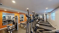 Located at 9951 Academy Rd in Philadelphia, PA, this fitness center offers a focused and functional layout across a single floor, comprising a welcoming entrance and a fully equipped exercise room. The entrance features warm wood-look flooring, white painted brick walls, and a glazed door that allows natural light to filter in, creating an inviting first impression. Signage near the entry provides facility information, setting a professional tone from the moment you walk in. The exercise room is finished with durable rubber flooring suited for high-traffic fitness use, and is illuminated by recessed ceiling lights throughout. The space is well-stocked with cardio equipment, including multiple treadmills and elliptical machines, as well as cable weight machines and a flat bench for strength training. A bold orange accent wall adds energy and visual interest to the room, while large wall-mounted mirrors amplify the sense of space and allow users to monitor their form. Windows along one wall bring in natural light, contributing to an open and airy atmosphere. A wall-mounted split AC unit ensures climate comfort year-round. The overall design is clean, practical, and purpose-built to support a comprehensive workout experience.